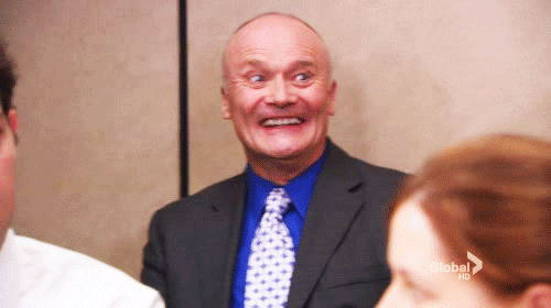 Creed Bratton from The Office in a dark suit and blue shirt with a wide, wild-eyed grin